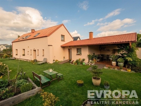 Zahrada u rodinn�ho domu s pe�liv� udr�ovan�m tr�vn�kem, zahradn� n�bytek a pergola. St�echa domu s �ervenou ta�kou, klidn� okol�.