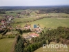 Leteck� pohled na malebnou vesnici obklopenou zelen�mi poli a lesy, s domy s �erven�mi st�echami a p�ilehlou p��rodou.